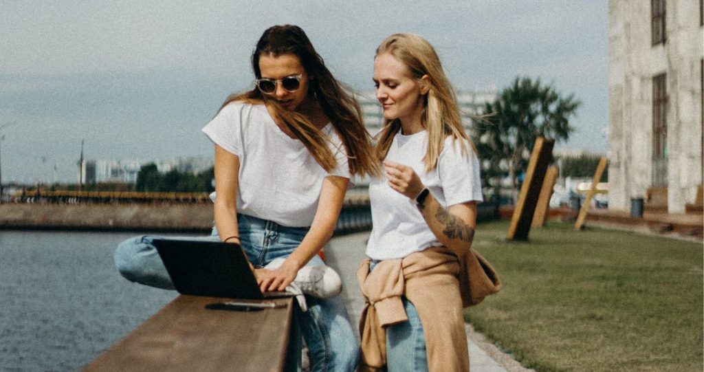 Na zdjęciu dwie kobiety. Kobieta siedząca na murku, ubrana jest w białą koszulkę, czarne okulary, niebieskie dżinsy i ma przed sobą laptopa. Stojąca obok niej kobieta w białej koszulce, jasnych dżinsach i beżowej bluzie zawiązanej na biodrach patrzy na laptopa.